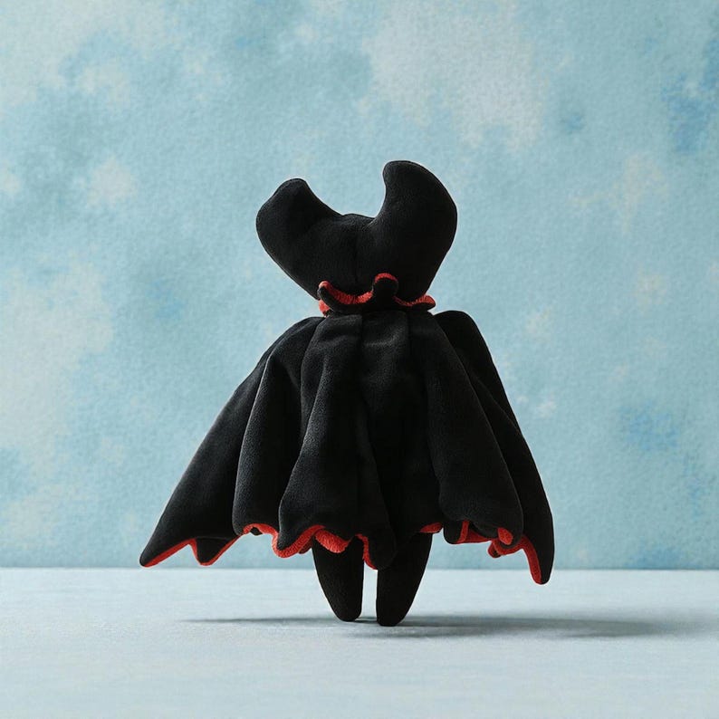 May include: A plush black bat-like figure with a red trim. The figure has a cape-like design with a ruffled red collar and two curved horns. The toy is set against a light blue background.