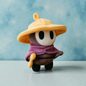 May include: A plush toy character with a yellow hat, purple cape, and a brown body. The toy has a white face with two black oval eyes and a small yellow accessory. The toy is standing on a light blue surface.