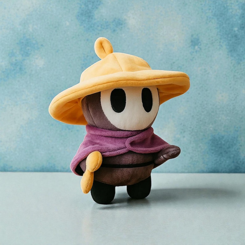 May include: A plush toy character with a yellow hat, purple cape, and a brown body. The toy has a white face with two black oval eyes and a small yellow accessory. The toy is standing on a light blue surface.