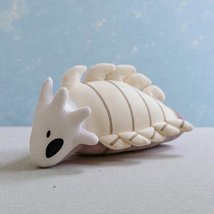May include: A plush toy shaped like a stylized sea creature. The toy has a white head with black eyes and a mouth, a cream-colored body with brown stripes, and cream-colored fins. The toy is on a light gray surface.