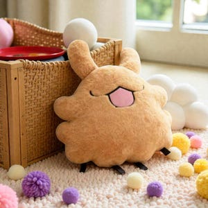 May include: A plush, tan-colored creature with large ears and a pink mouth. The plush toy is on a white rug with various colorful pom-poms. A woven basket and other decorative items are in the background.