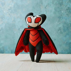 May include: A plush toy of a character with a black head and limbs, a white face with red eyes, and a red and black body. The character has a red cape. The toy is standing against a blue background.