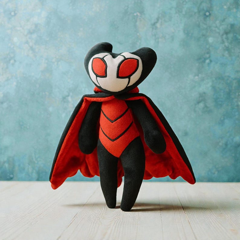 May include: A plush toy of a character with a black head and limbs, a white face with red eyes, and a red and black body. The character has a red cape. The toy is standing against a blue background.
