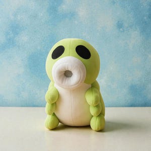 May include: A plush toy in the shape of a green creature with a large white mouth and two black eyes. The toy has small, rounded arms and legs. The background is a light blue with white accents.
