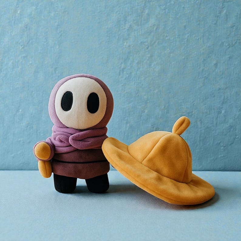May include: A plush toy of a Shy Guy character from the Super Mario video game series. The figure is purple, with a white mask and black eye holes. A yellow hat rests beside it.