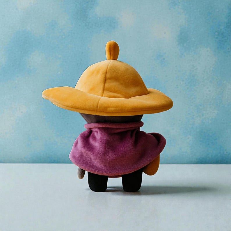 May include: A plush doll with a yellow hat and a purple cape. The doll has black feet and a brown body. The hat has a small, rounded top. The cape is draped over the doll's shoulders. The background is a light blue color.