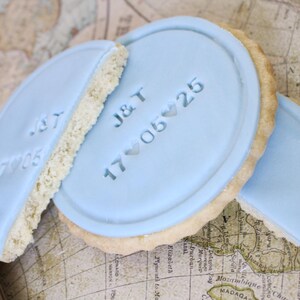 Peut inclure: Des biscuits glac&eacute;s bleu clair, avec les initiales "J&T" et la date "17 05 25", sont pos&eacute;s sur une carte vintage. Les biscuits sont ronds avec un bord clair et textur&eacute;. Un biscuit est cass&eacute; en deux.