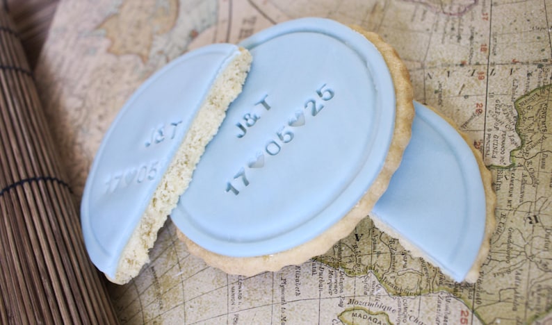 Peut inclure: Des biscuits glac&eacute;s bleu clair, avec les initiales "J&T" et la date "17 05 25", sont pos&eacute;s sur une carte vintage. Les biscuits sont ronds avec un bord clair et textur&eacute;. Un biscuit est cass&eacute; en deux.