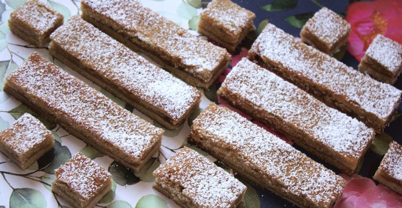 May include: A selection of rectangular and square pastries, dusted with icing sugar, are arranged on a floral patterned surface. The pastries are a light brown colour and appear layered. The background has green and blue tones.