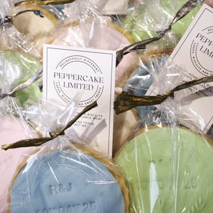 Peut inclure: Gros plan sur plusieurs biscuits emball&eacute;s individuellement dans du cellophane transparent. Chaque biscuit a un gla&ccedil;age de couleur pastel diff&eacute;rente et est nou&eacute; avec un ruban dor&eacute;. Les biscuits ont des motifs et du texte en relief. L'emballage comprend une carte "PepperCake Limited".