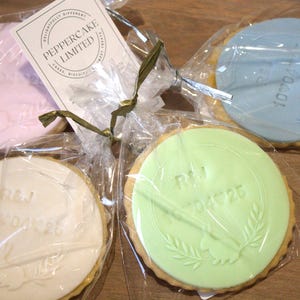Peut inclure: Quatre biscuits ronds, chacun emball&eacute; individuellement dans du cellophane transparent. Les biscuits ont un gla&ccedil;age de couleur pastel avec des motifs et du texte en relief. Un biscuit est rose, un est bleu, un est vert et un est blanc.