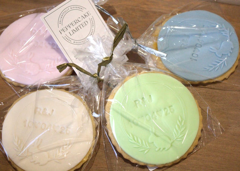 Peut inclure: Quatre biscuits ronds, chacun emball&eacute; individuellement dans du cellophane transparent. Les biscuits ont un gla&ccedil;age de couleur pastel avec des motifs et du texte en relief. Un biscuit est rose, un est bleu, un est vert et un est blanc.