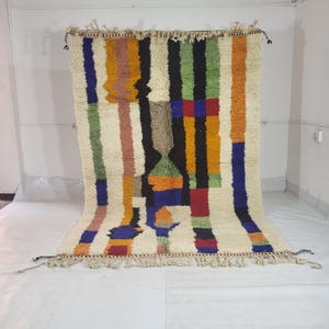 May include: A rectangular, hand-woven rug with a cream-colored background and vertical stripes in various colors, including blue, orange, green, and black. The rug has a geometric design with a fringed edge and tassels at the corners.