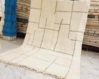 May include: A large, off-white rug with a geometric design of gray lines. The rug has a thick, textured pile and a fringe along the bottom edge. The rug is displayed on a marble floor, with other rugs stacked in the background.