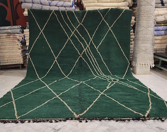 May include: A large, dark green rug with a diamond pattern in a lighter beige color. The rug has a fringe along the bottom edge. The rug is displayed on a marble floor, with other rugs and textiles in the background.
