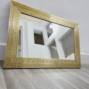 Puede incluir: Espejo rectangular dorado con un marco ornamentado y tallado. El marco presenta patrones y diseños detallados. El espejo refleja una pared blanca con detalles arquitectónicos y un suelo gris.