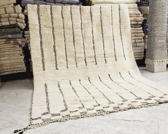 May include: A large, off-white rug with vertical black stripes. The rug has a textured surface and a fringed edge. Stacks of other rugs are visible in the background, suggesting a rug shop or display.