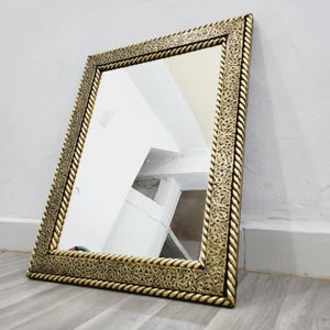 Puede incluir: Un espejo rectangular con un marco dorado ornamentado. El marco presenta un patrón detallado y un borde en forma de cuerda. El espejo está apoyado contra una pared gris claro y un suelo de madera.