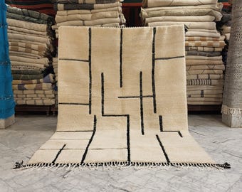 May include: A cream-colored rug with a geometric black line design. The rug has a thick pile and a fringe along the bottom edge. The rug is displayed in a shop with stacks of other rugs in the background.