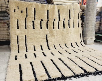 May include: A large, cream-colored rug with a geometric pattern of vertical black lines. The rug has a thick, textured pile and a black fringe along the bottom edge. The rug is displayed in a shop setting, with other rugs and textiles visible in the background.