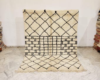 May include: Cream-colored rug with a geometric design. The rug features a pattern of black diamonds and rectangular shapes. The rug has a fringe along the bottom edge and is displayed against a white wall.