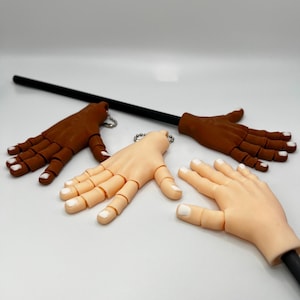 May include: Articulated hand props in various skin tones, including light and dark brown. Each hand has a white nail color and is attached to a black rod with a keychain. The hands are designed for posing and display.