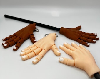 Peut inclure: Des accessoires de mains articulées de différentes couleurs de peau, dont le marron clair et foncé. Chaque main a des ongles blancs et est fixée à une tige noire avec un porte-clés. Les mains sont conçues pour la pose et l'exposition.