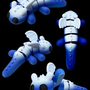 Op de afbeelding: Verschillende weergaven van een blauw en wit gearticuleerd speelgoeddraak. Het speelgoed heeft een gesegmenteerd lichaam, afgeronde ledematen en vleugelachtige structuren. De draken hebben zwarte vlekken op een zwarte achtergrond.