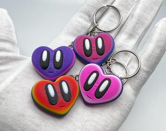 Peut inclure: Quatre porte-clés en forme de cœur, violets, roses, rouges et bleus, chacun avec une paire d'yeux ovales blancs et un contour noir. Les porte-clés sont posés sur une surface blanche.