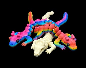 Peut inclure: Quatre figurines de lézard articulées. Trois sont aux couleurs vives de l'arc-en-ciel, et une est blanc cassé. Les lézards ont des corps segmentés et sont posés sur un fond noir. Les lézards arc-en-ciel ont des couleurs rose, bleu, jaune et orange.