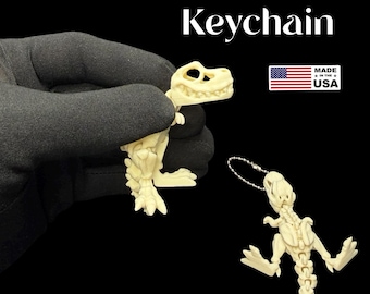 Peut inclure: Porte-clés Mini T. Rex de couleur blanc cassé. Le porte-clés est un squelette de dinosaure miniature avec un porte-clés attaché. Le texte "Mini T. Rex Keychain" est affiché au-dessus du dinosaure. Le drapeau américain et "Made in the USA" sont également visibles.