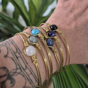 Könnte beinhalten: Eine Sammlung goldfarbener Armreifen, jeder mit einem anderen Edelstein: Türkis, Grau, Weiß, Blau, Lila und Schwarz. Die Edelsteine sind mit Draht auf die Armreifen gewickelt. Der Hintergrund ist grünes Laub.