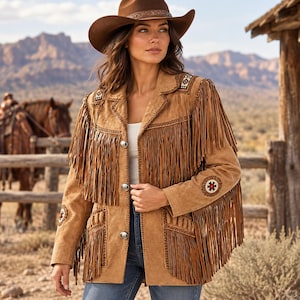 May include: A tan suede jacket featuring long fringe detailing on the chest and sleeves, silver-toned buttons, and decorative circular embroidery on the shoulders and forearms. The jacket is worn over a white top and blue denim jeans.