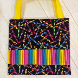 May include: A square fabric tote bag with bright yellow nylon handles. The main body features a black background with a scattered pattern of colorful crayons. A horizontal band across the center displays a vertical rainbow stripe crayon design.