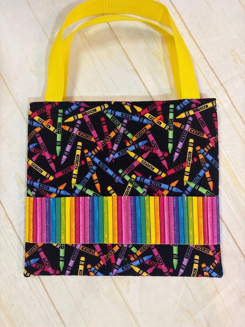 May include: A square fabric tote bag with bright yellow nylon handles. The main body features a black background with a scattered pattern of colorful crayons. A horizontal band across the center displays a vertical rainbow stripe crayon design.