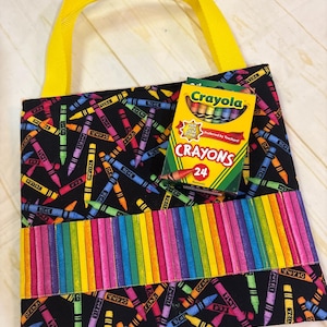 May include: A black fabric tote bag with a colorful crayon print, featuring bright yellow handles and a horizontal pocket with a rainbow-striped pattern. A box of 24 Crayola crayons is placed on the front of the bag, displaying the brand name.