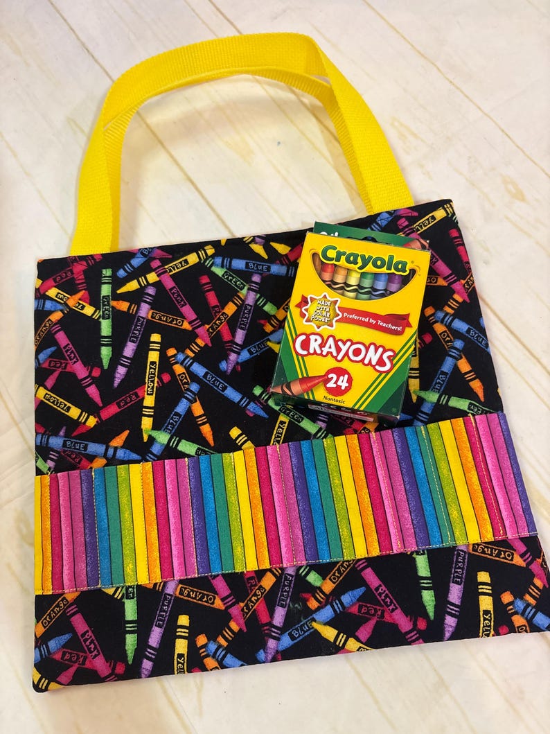 May include: A black fabric tote bag with a colorful crayon print, featuring bright yellow handles and a horizontal pocket with a rainbow-striped pattern. A box of 24 Crayola crayons is placed on the front of the bag, displaying the brand name.