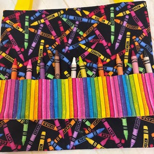 May include: A black fabric bag featuring a colorful crayon print with yellow straps. The bag includes a front pocket with vertical slots containing individual crayons in various colors, including orange, pink, purple, brown, white, yellow, and red.