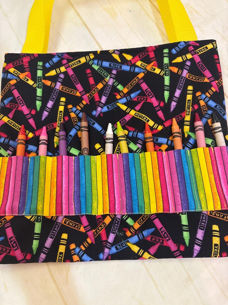 May include: A black fabric bag featuring a colorful crayon print with yellow straps. The bag includes a front pocket with vertical slots containing individual crayons in various colors, including orange, pink, purple, brown, white, yellow, and red.
