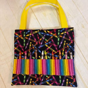 May include: A black fabric tote bag featuring a colorful crayon print pattern. The bag includes bright yellow ribbon handles and a horizontal front pocket section designed with vertical rainbow stripes, currently holding ten individual wax crayons.
