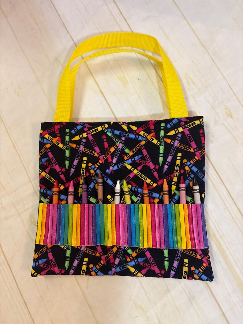 May include: A black fabric tote bag featuring a colorful crayon print pattern. The bag includes bright yellow ribbon handles and a horizontal front pocket section designed with vertical rainbow stripes, currently holding ten individual wax crayons.