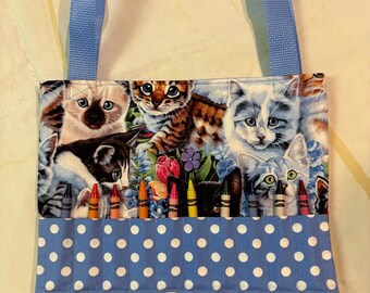 May include: A blue tote bag with a cat print and a blue handle. The bag has a blue pocket with white polka dots and holds several crayons. The cat print features various cat breeds and floral accents.