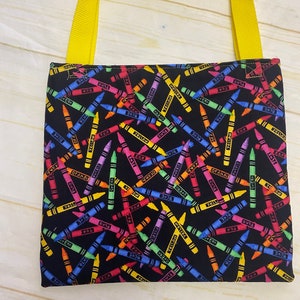 May include: A square fabric tote bag featuring a black background with a repeating pattern of colorful crayons in shades of red, blue, yellow, green, orange, and purple. The bag includes two bright yellow woven fabric handles attached at the top.