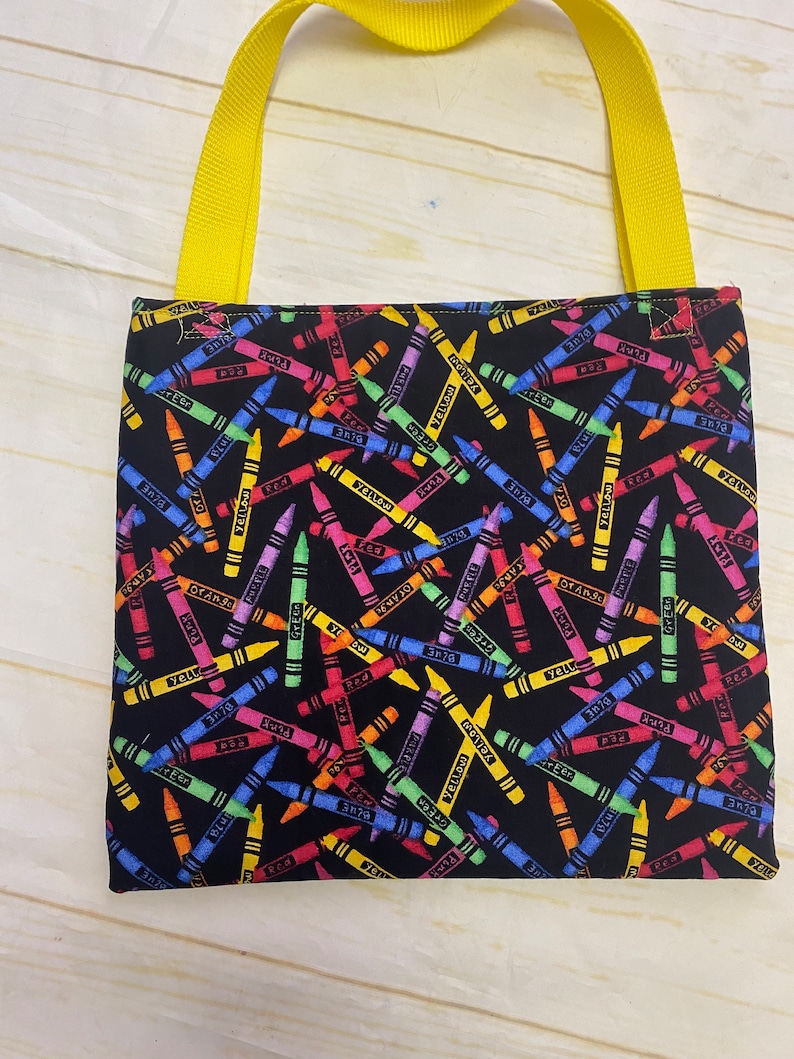 May include: A square fabric tote bag featuring a black background with a repeating pattern of colorful crayons in shades of red, blue, yellow, green, orange, and purple. The bag includes two bright yellow woven fabric handles attached at the top.
