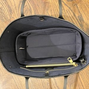 May include: Black tote bag with a navy blue interior and a matching organizer insert. The bag has black handles and a gold zipper. The organizer has multiple compartments for storage. The bag is open, showing the interior.