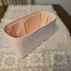 May include: A light pink rectangular felt bag organizer insert with rounded corners and multiple interior compartments. The structure is soft yet maintains its shape, featuring stitched seams along the top edges and internal dividers for organization.
