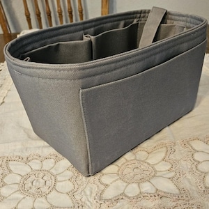 May include: A rectangular gray fabric bag organizer insert featuring a structured design with multiple interior compartments, open side pockets, and a central vertical strap, constructed from a durable woven material with reinforced stitched edges.