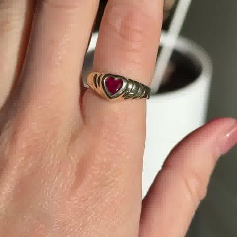 Pu&ograve; includere: Un anello color oro con una pietra rossa centrale a forma di cuore. La fascia presenta un design a coste e testurizzato su entrambi i lati dell'incastonatura a cuore, creando un motivo distinto che avvolge il dito con una finitura metallica lucida.