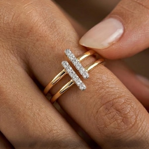 Könnte beinhalten: Ein goldener Ring mit zwei parallelen Bändern und zwei vertikalen Stäben, die mit kleinen, funkelnden weißen Steinen besetzt sind. Der Ring wird an einem Finger gezeigt, was sein elegantes Design und den Kontrast zwischen Gold und weißen Steinen hervorhebt.