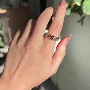 Pu&ograve; includere: Un anello in oro con una pietra preziosa rossa a forma di cuore al centro. La fascia dell'anello presenta una consistenza a coste o scanalata che si estende attorno al dito. Il metallo ha una finitura lucida e la pietra rossa &egrave; ben incastonata.